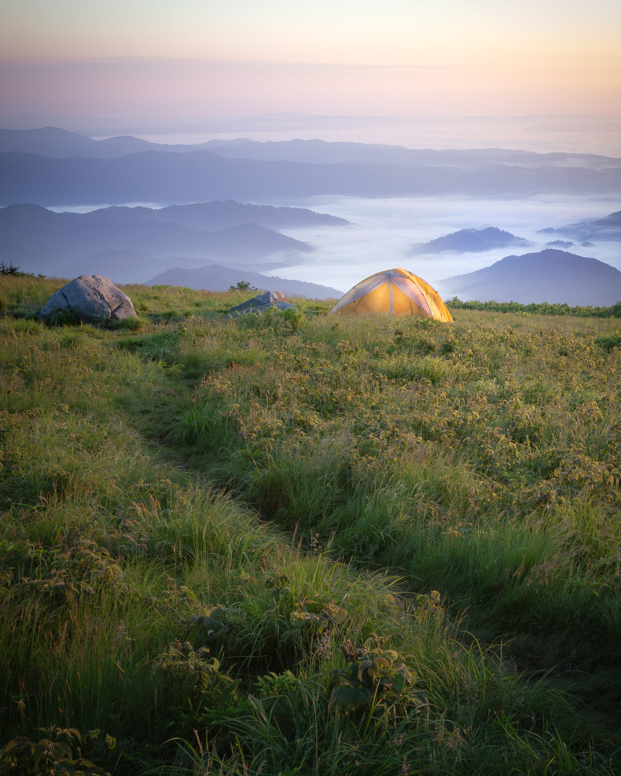 A glowing tent on an open grassy bald at sunrise in the Roan Highlands, with layered blue ridges fading into the distance.